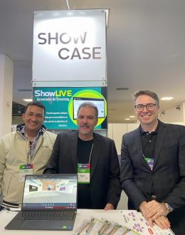 Atrás de uma bancada com notebook aberto, da esquerda para a direita, um homem de pele negra e cabelos pretos, um homem de pele branca, cabelos e cavanhaque grisalho, um homem de cabelo grisalho e óculos de grau sorriem.