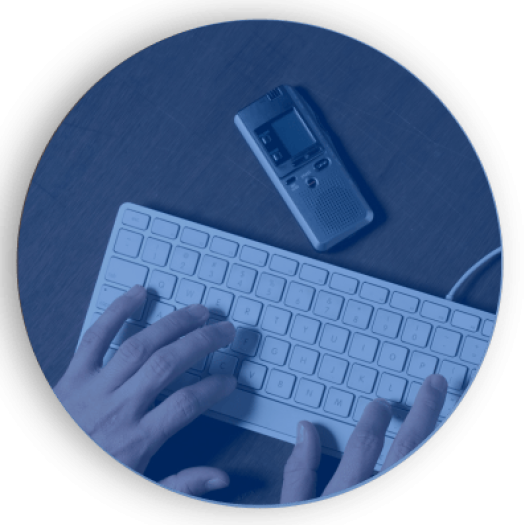 Foto azulada. Em uma mesa, uma pessoa de pele branca com as mãos em um teclado branco. Acima do teclado, um gravador de voz.